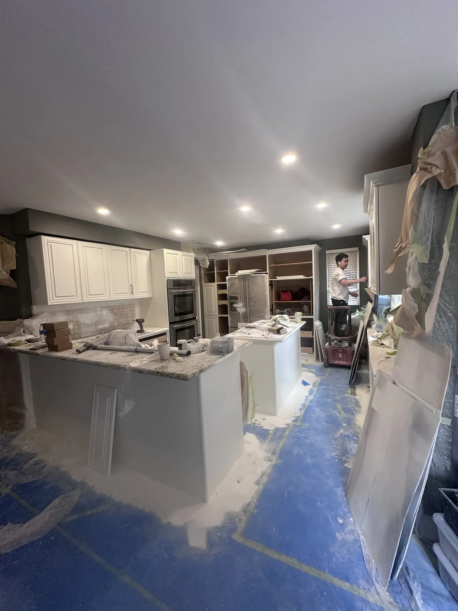 The kitchen mid-job with the floor covered in blue protection sheet, cabinetry masked and primed, and a sprayer working in the room