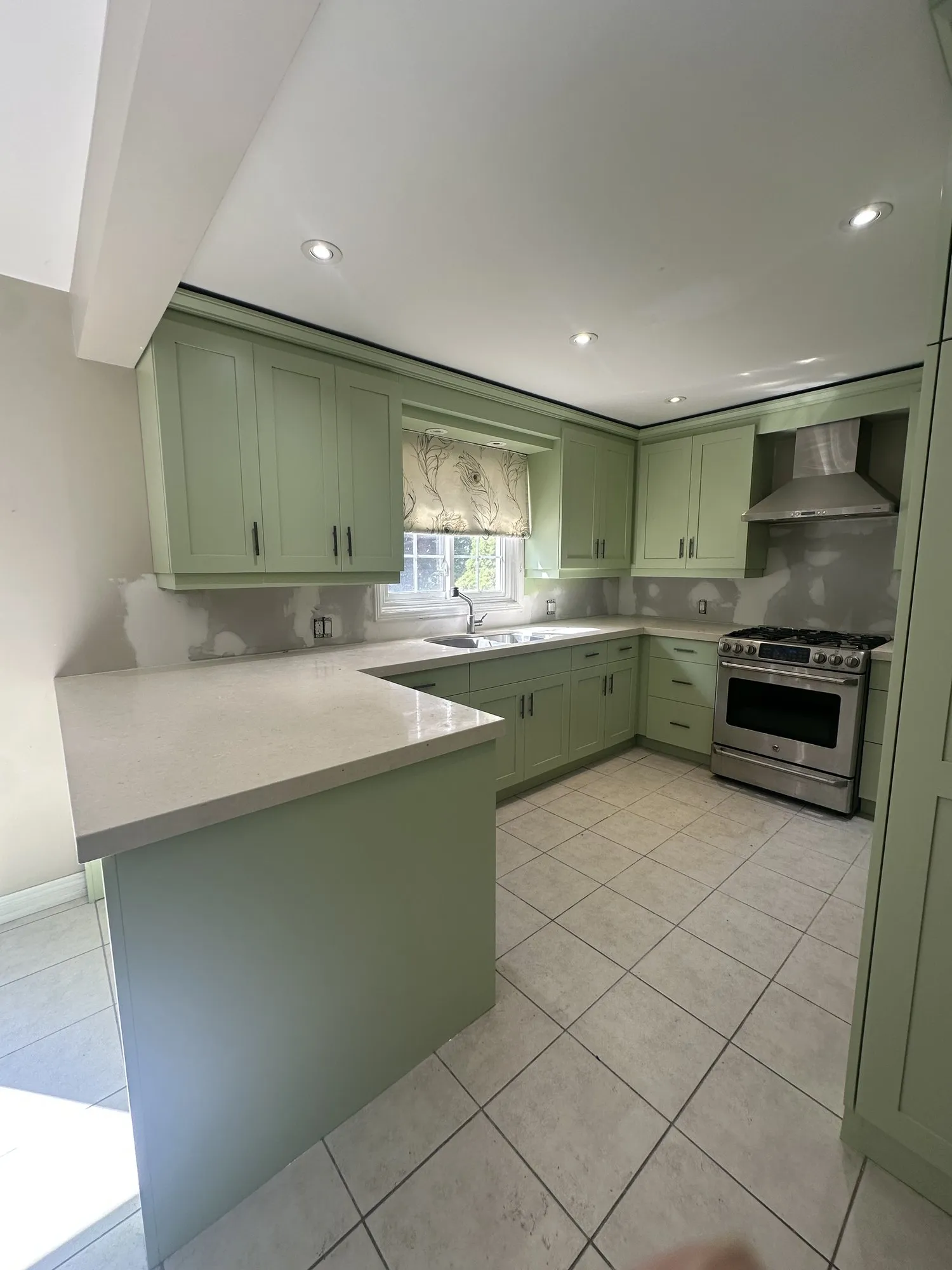 The finished kitchen seen past the peninsula, the light green cabinetry in place and the wall behind the counters stripped back to bare patched drywall where the old mosaic tile was