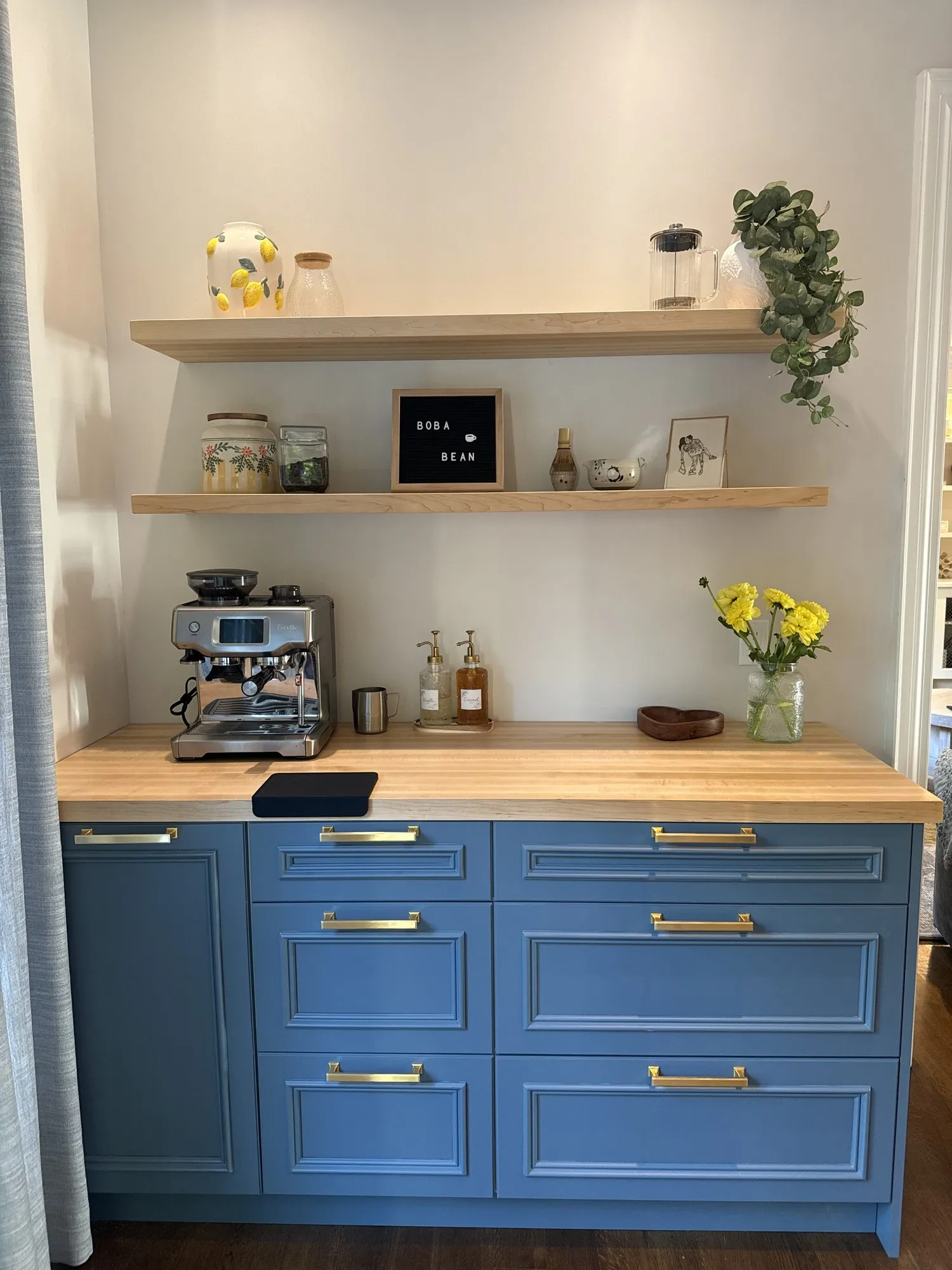 The finished coffee station in use, blue drawers with brass bar pulls under a butcher-block counter, espresso machine and jars on top and two wood shelves above