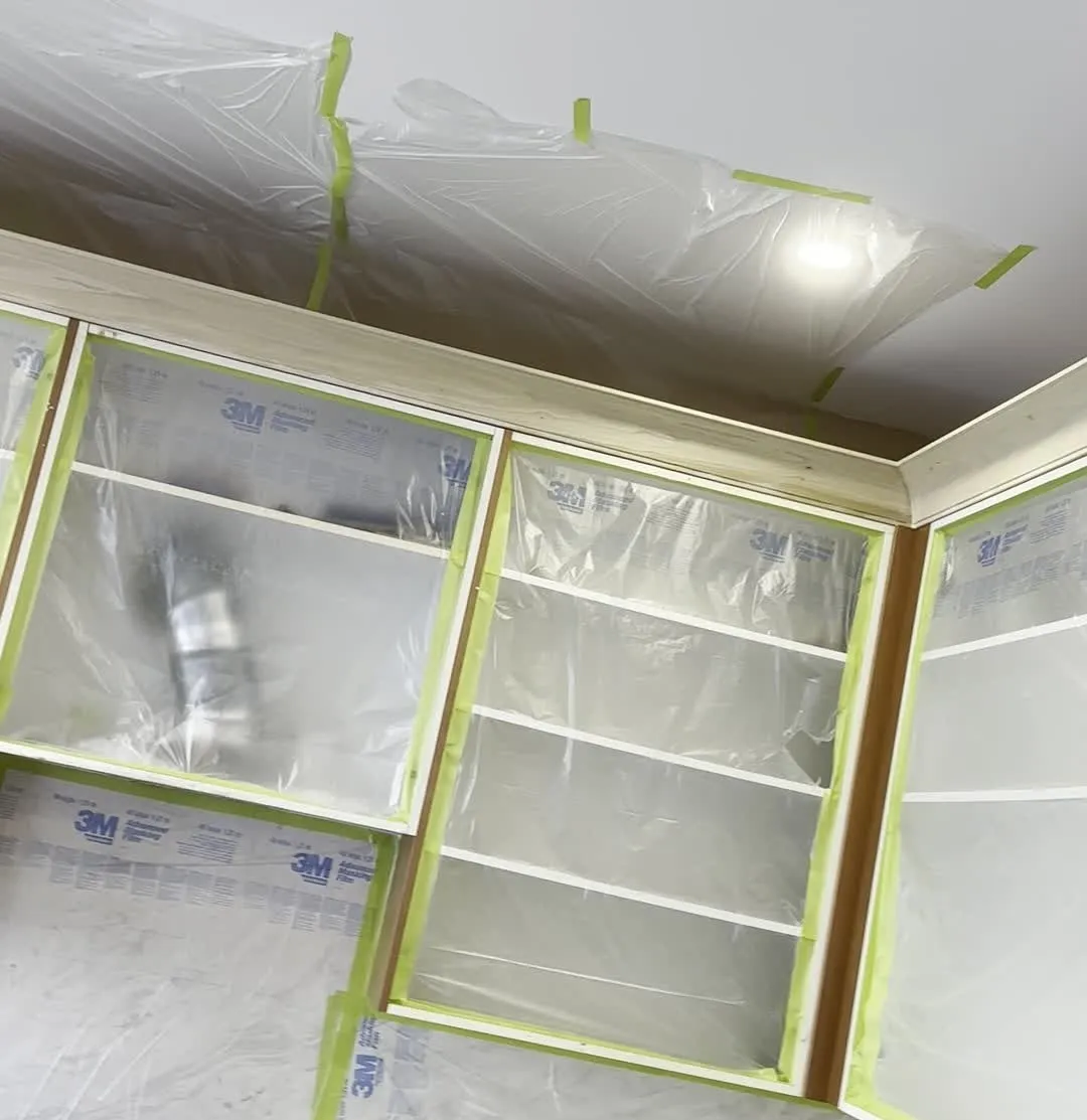 New bare wood crown moulding installed along the top of the upper cabinets, with every cabinet opening sealed in plastic film and green tape ready for spraying