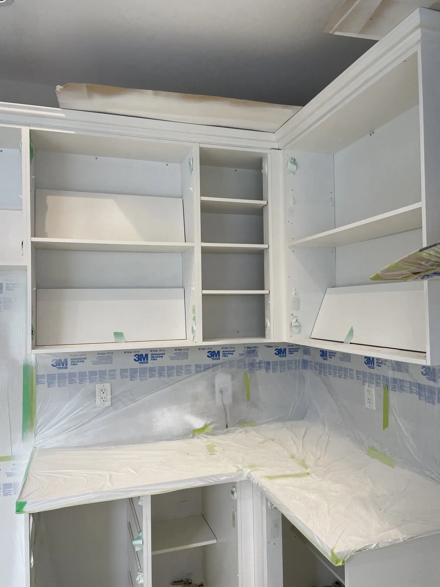 The kitchen on spray day with every cabinet door removed, the interiors sealed behind masking film and the counters and floor fully sheeted