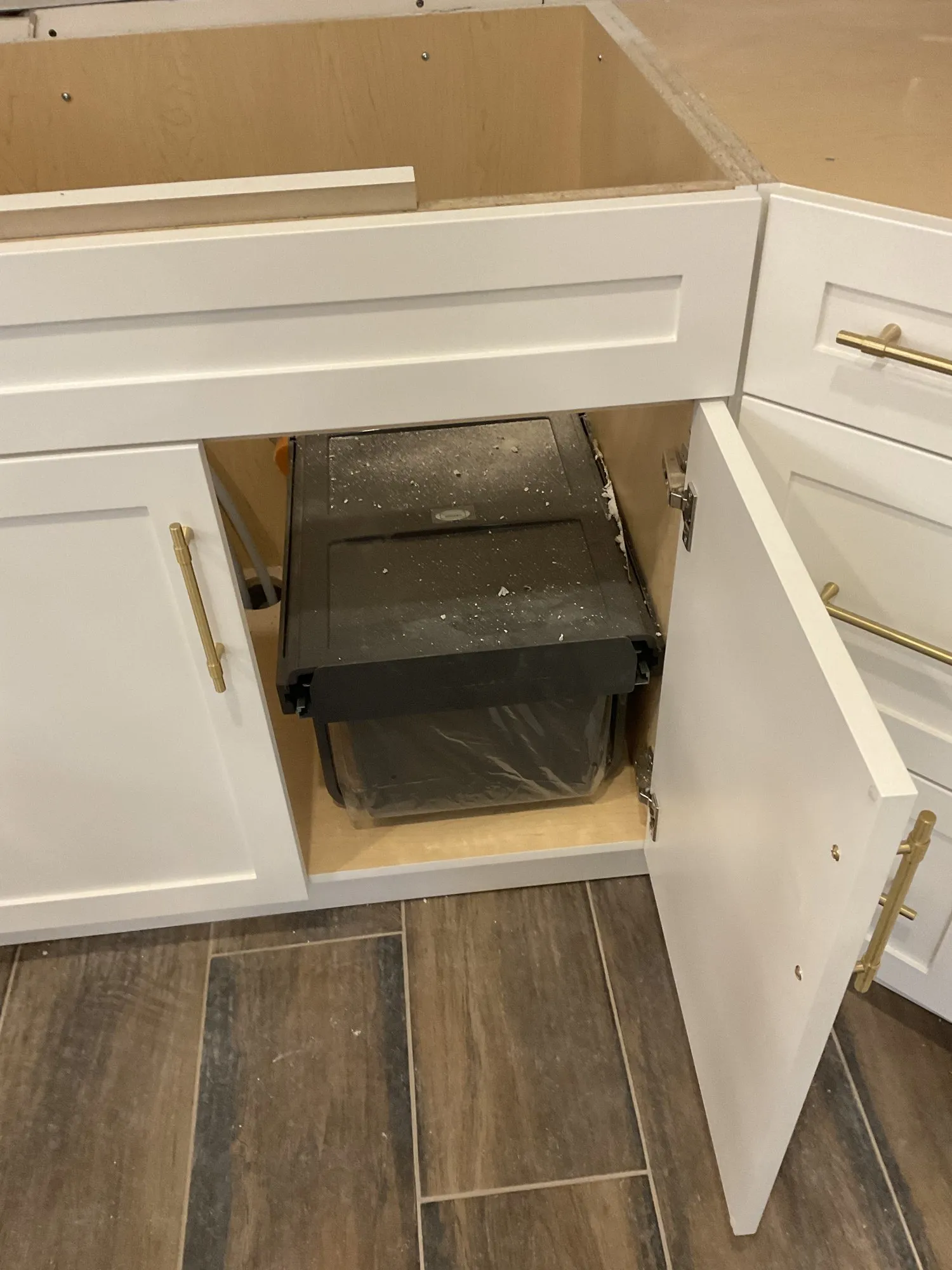 The under-sink cabinet standing open, showing the waste bin on its pullout runner behind a white shaker door with a brass handle