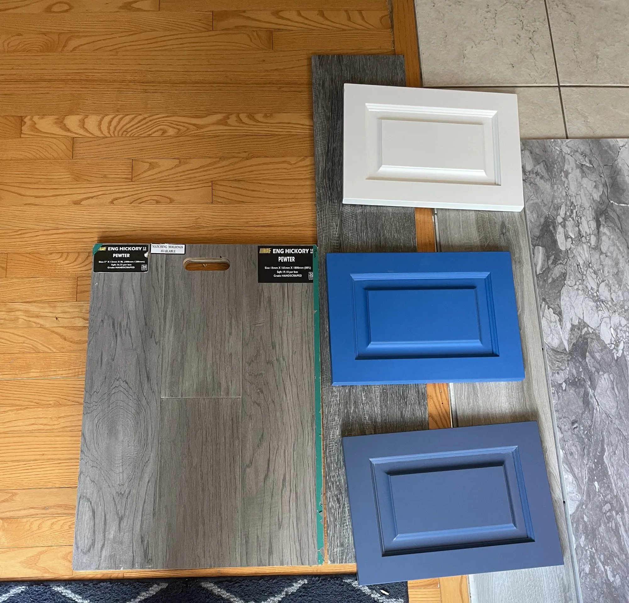 A white door sample, two blue door samples, a grey-veined stone slab and engineered hickory flooring boards laid out together on the floor of the house