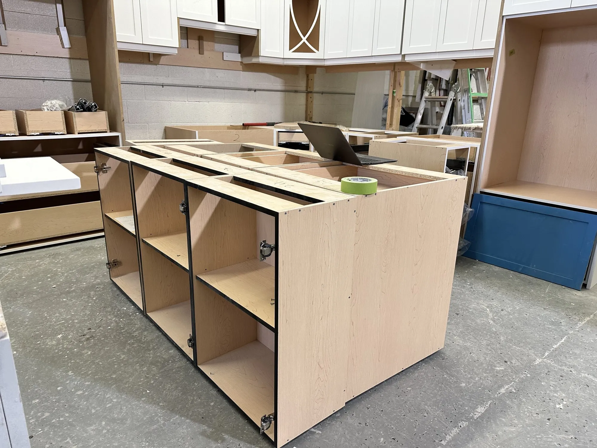 The island carcass part built on the shop floor, its plywood compartments open, with the finished blue panels and white mullion-glass doors stacked behind it