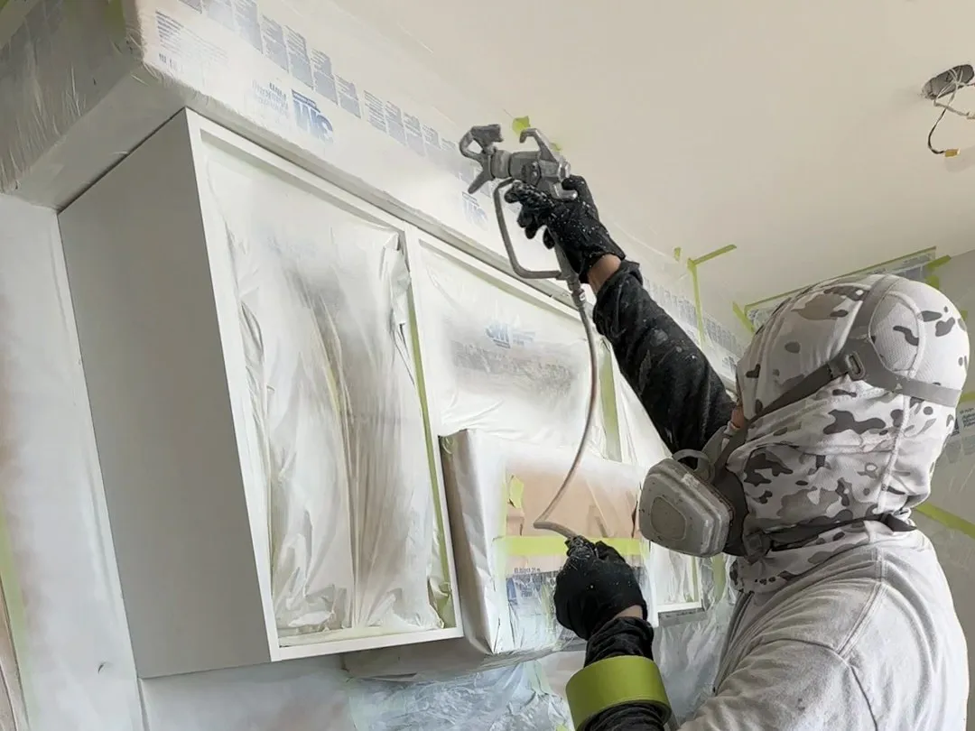 A sprayer in a hood and respirator spraying 2K polyurethane onto cabinet frames still fixed to the wall, the openings masked and the room sheeted behind him