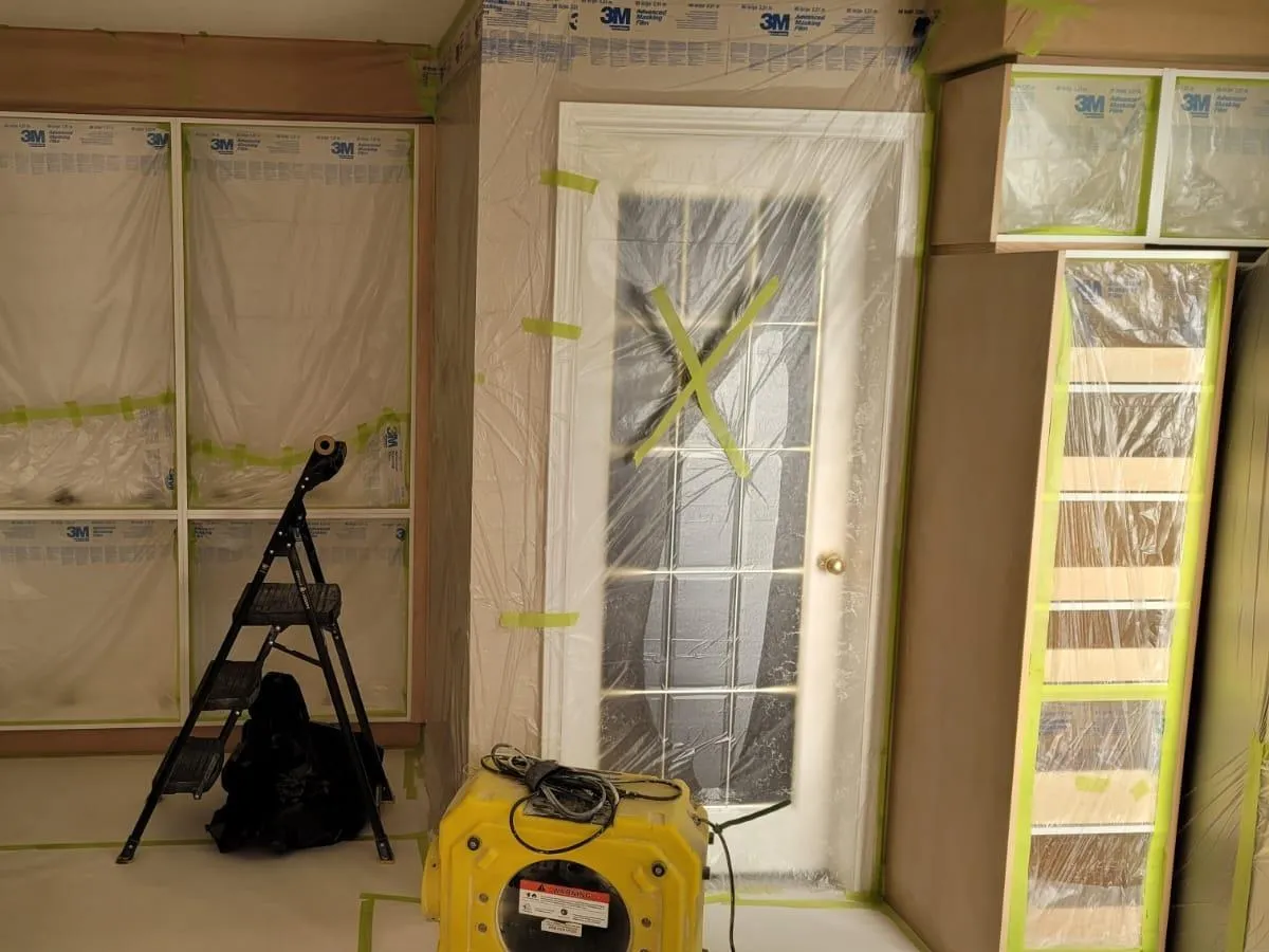 A room sealed before spraying — windows and door masked in sheeting and tape, the floor covered, the cabinets wrapped, and an air scrubber running on the floor