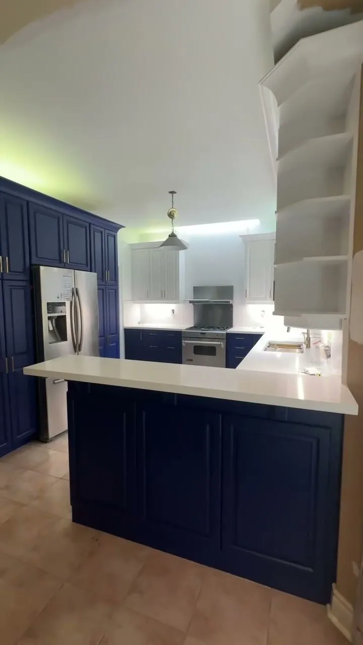 The finished Merton kitchen — Blue Grotto base cabinets and a peninsula under white quartz, Chantilly Lace uppers with glass doors, brushed brass pulls throughout