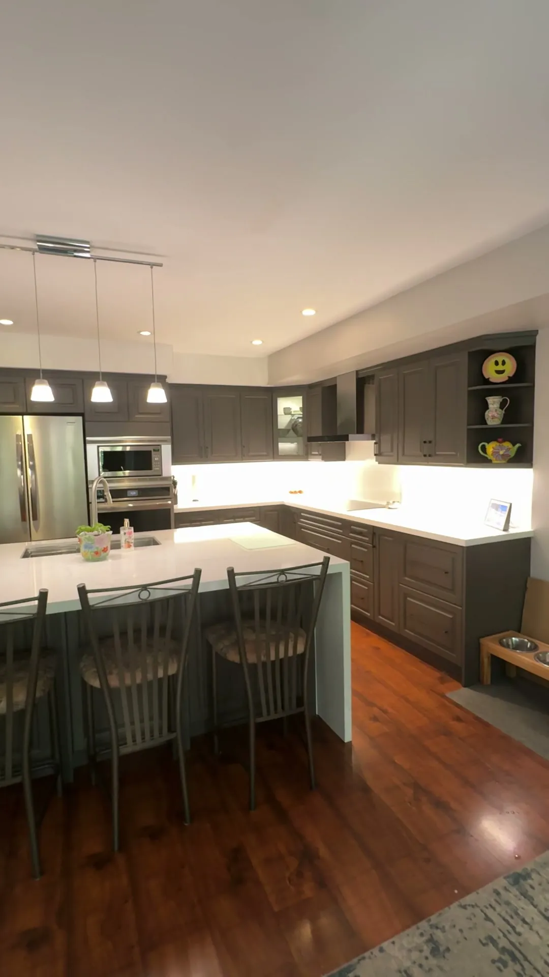 The finished Uptown Core kitchen — charcoal sprayed cabinets around a white quartz island with four stools, glass-front uppers lit from within, opening onto the living room