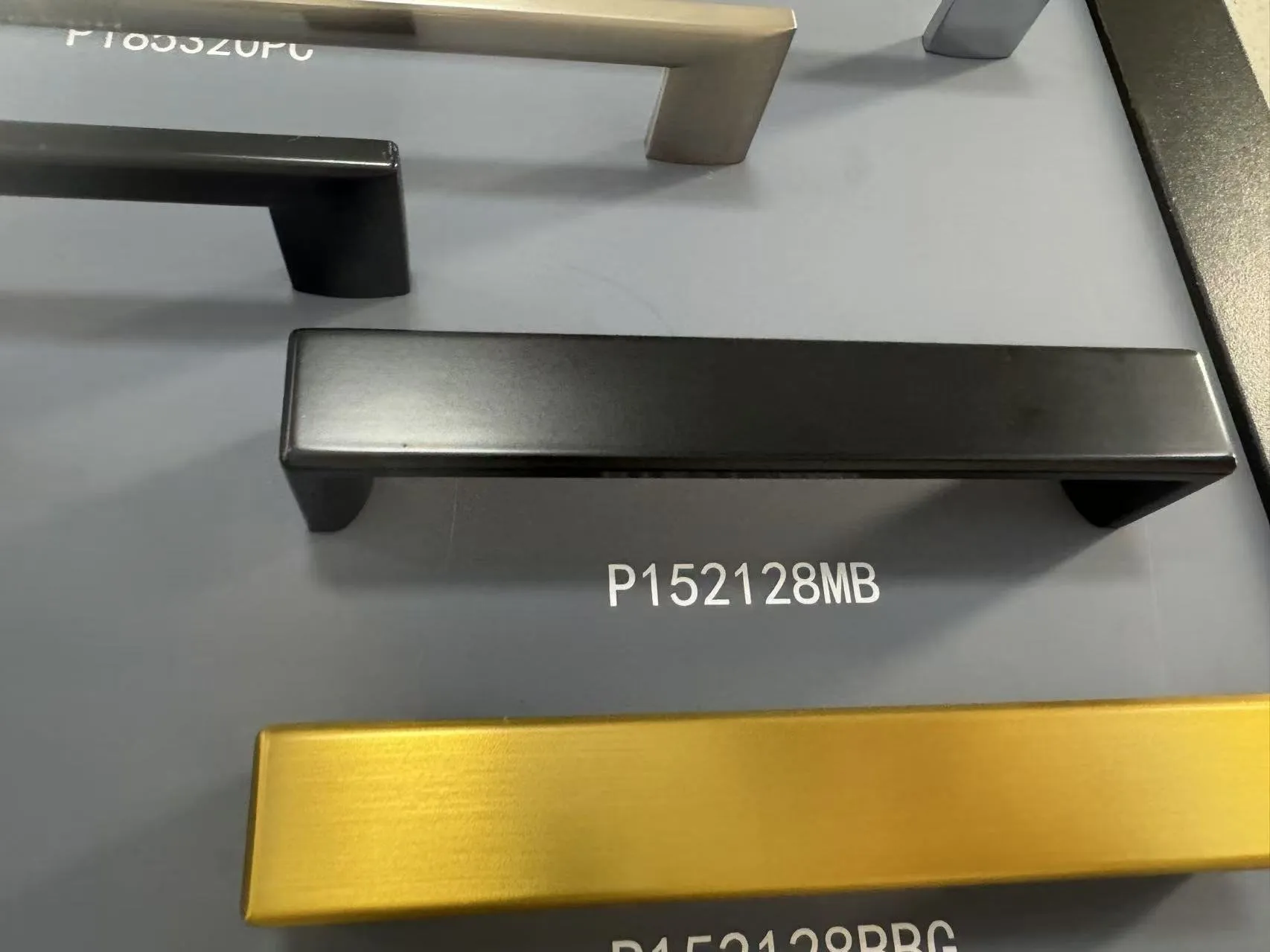 A matte black bar pull on a handle sample board, labelled P152128MB, beside a brushed gold pull