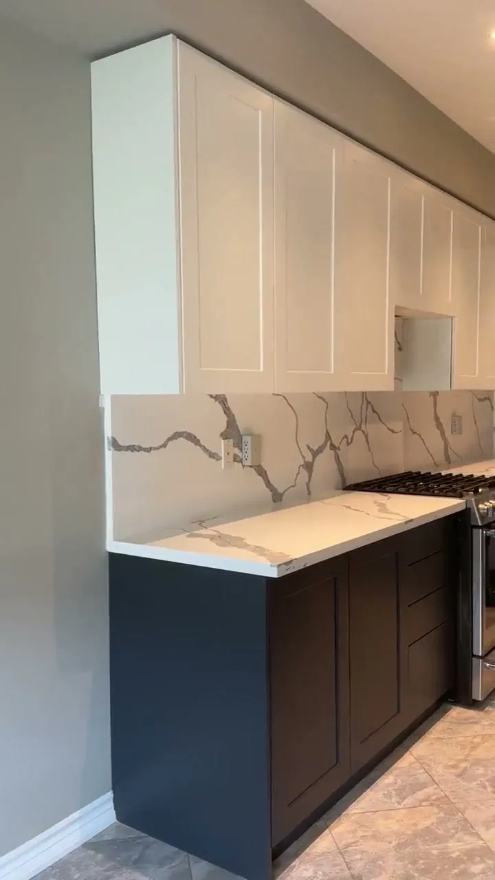 The finished Bronte kitchen — white shaker uppers over charcoal bases, quartz counters with the veining carried up the wall as a slab backsplash, and the original columns kept