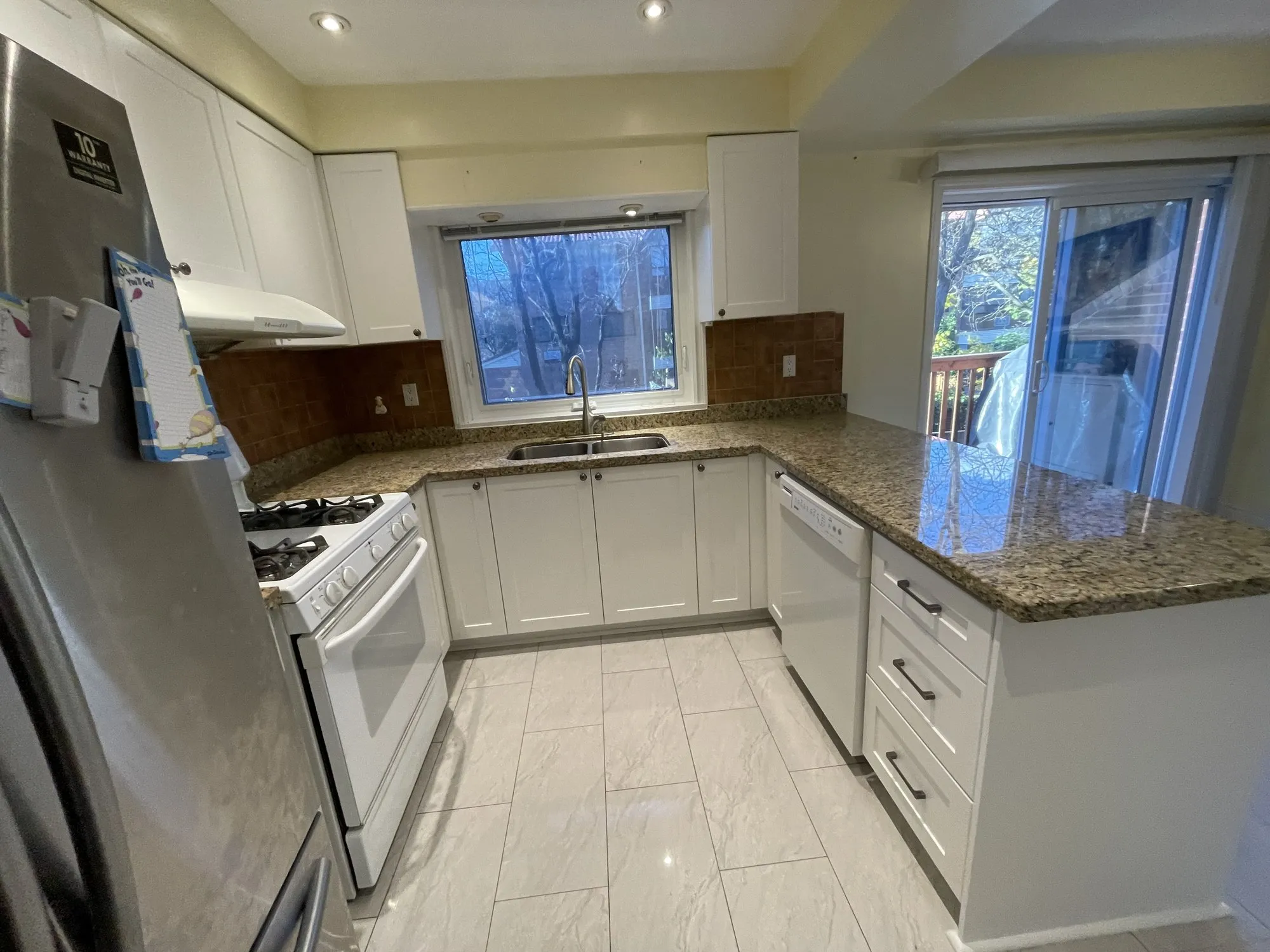 The same kitchen after refacing, with white MDF shaker doors, long dark bar handles, and the original granite counter and brick backsplash unchanged