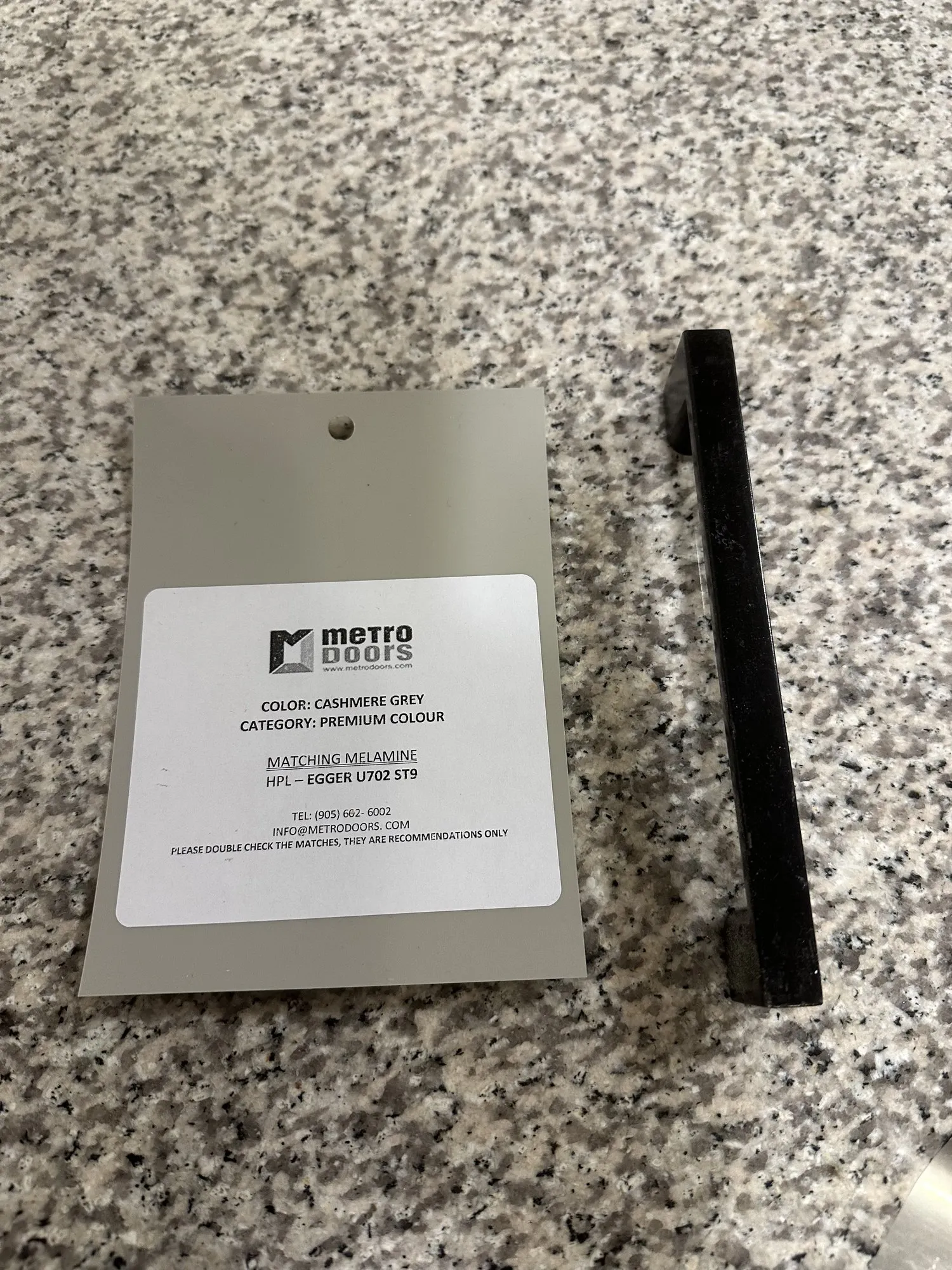 A Cashmere Grey door sample and a black bar handle laid on the client's own granite counter, the colour label visible on the sample