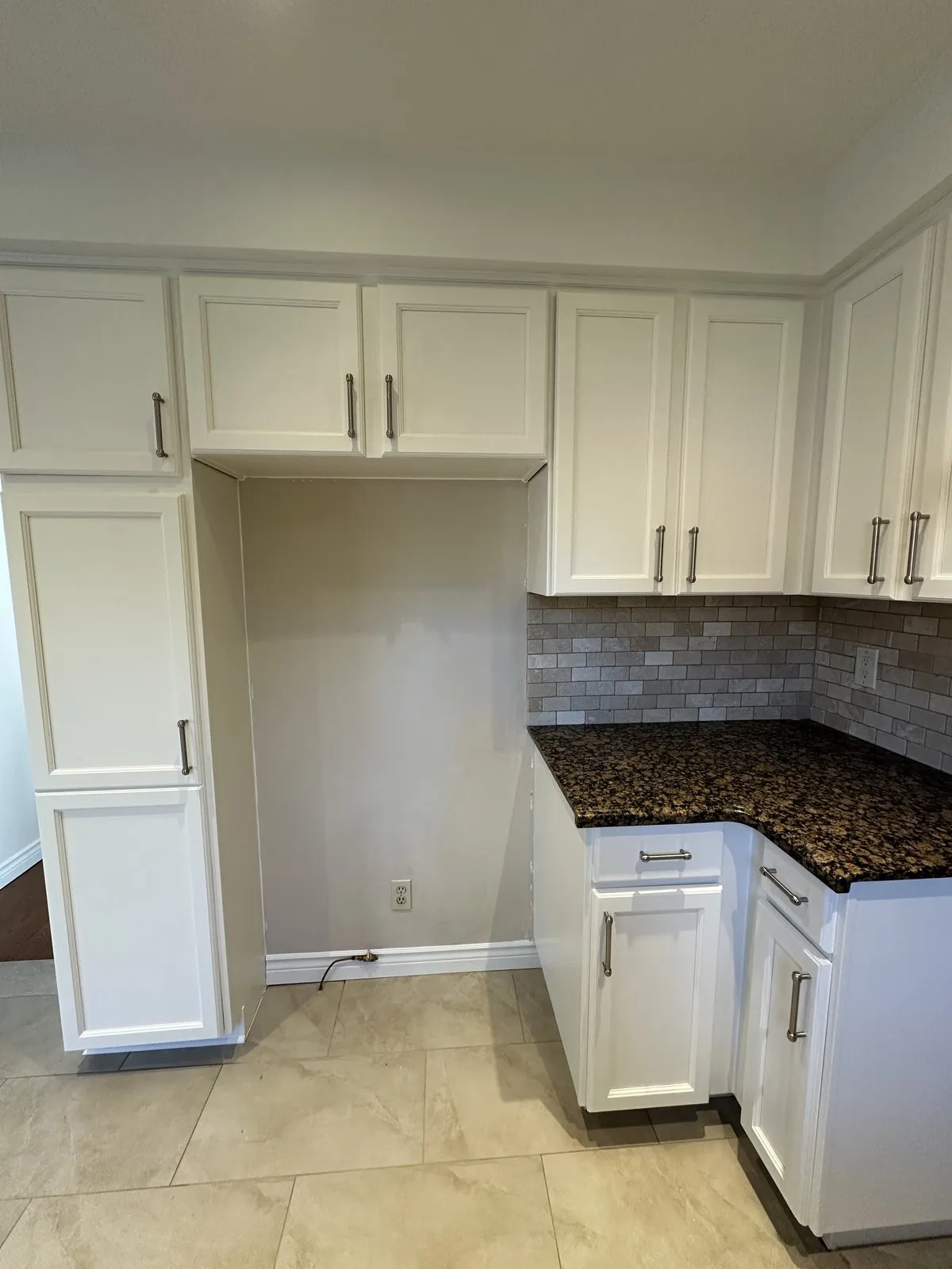Tall white pantry cabinet and white Shaker uppers around an open fridge space, beside a granite corner
