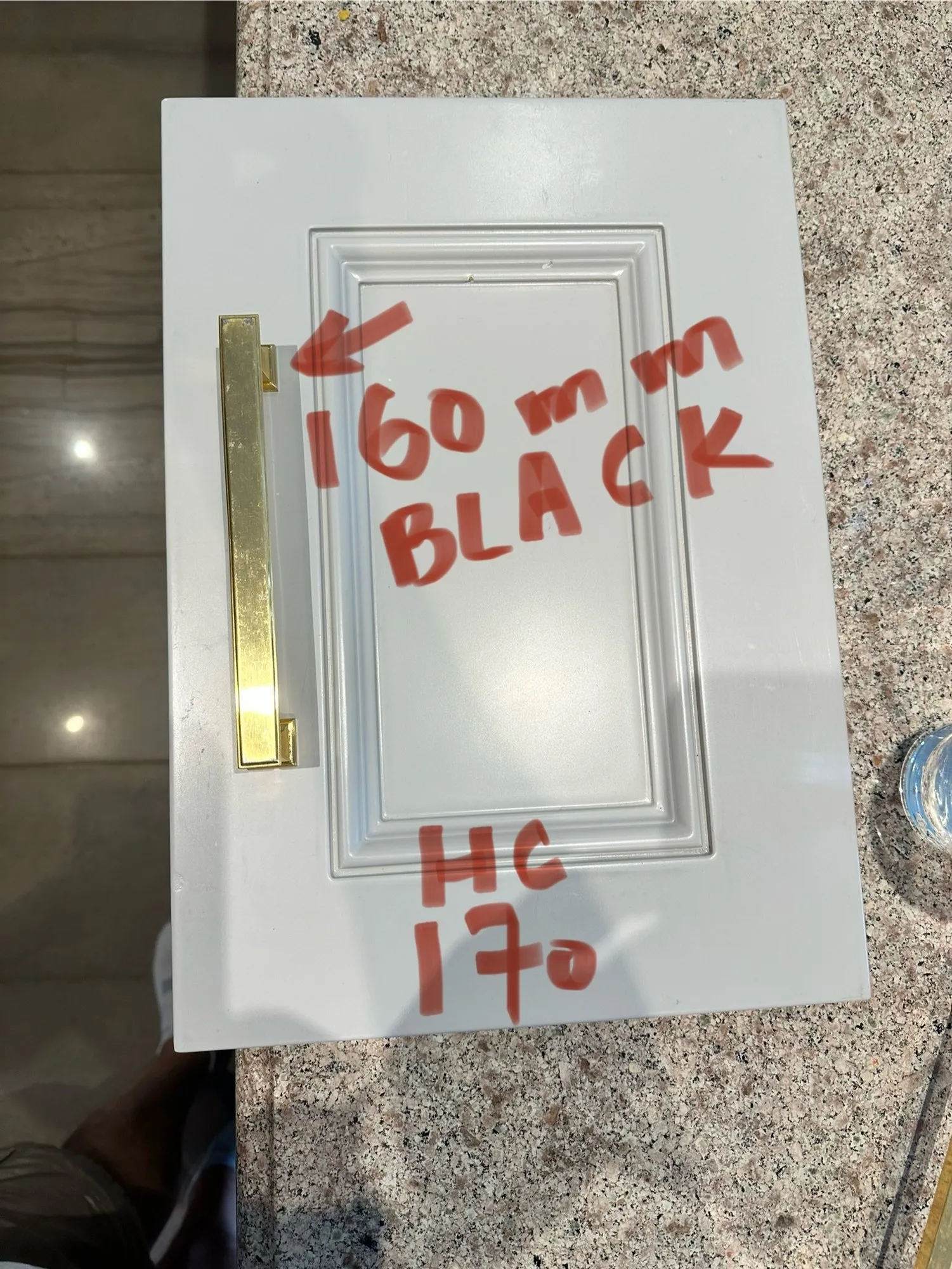 A white sample door with the handle specification written across it in red marker — 160 millimetre centres, black — beside a brass handle laid on the counter for comparison