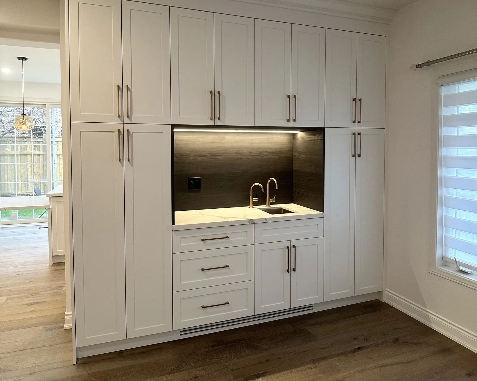 A full-height stone splashback in a dark wood-grain finish running behind a marble-veined countertop, with twin brass gooseneck taps at the sink