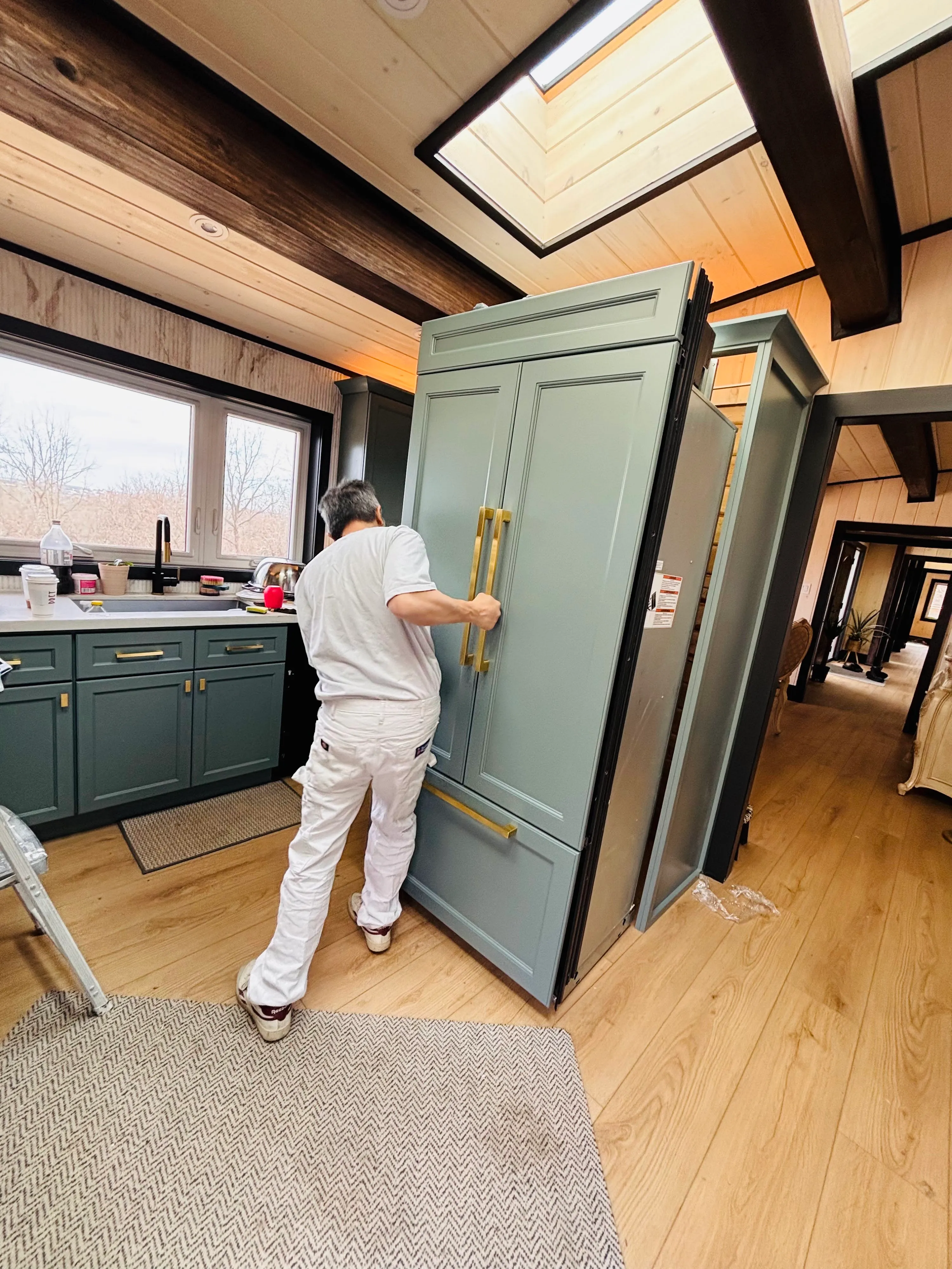 A technician fitting a custom sage-green panel with brass pulls to a panel-ready built-in fridge, matching the surrounding cabinetry exactly