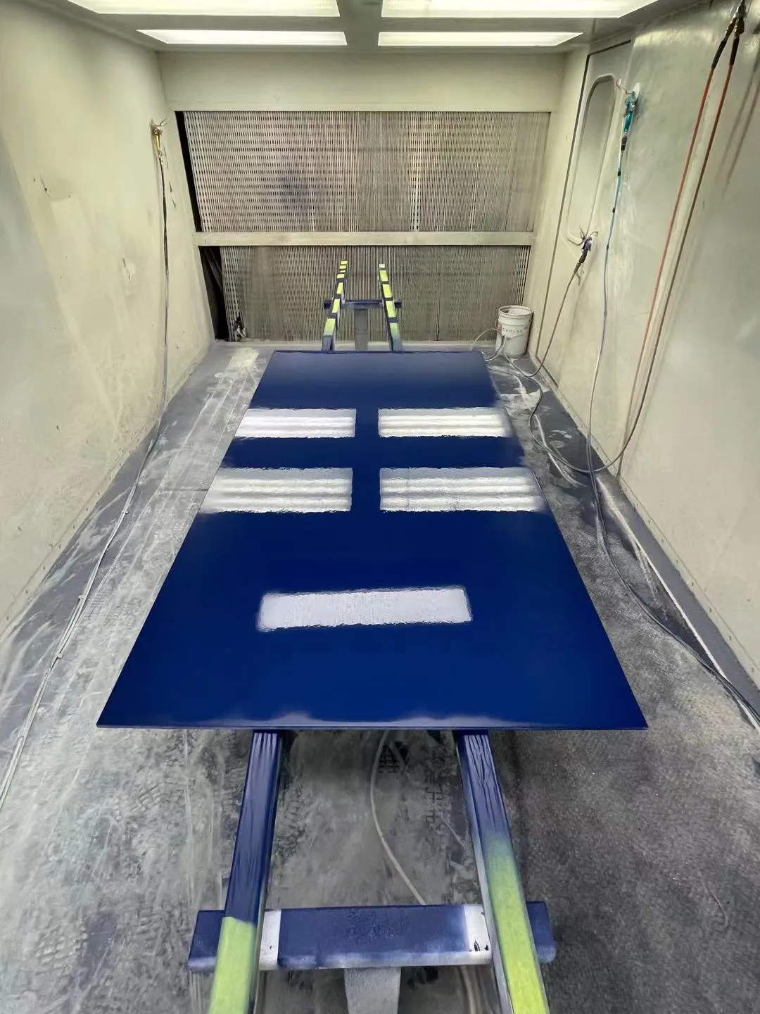 A cabinet panel sprayed glossy navy blue, laid flat on stands inside the spray booth, filter wall behind it