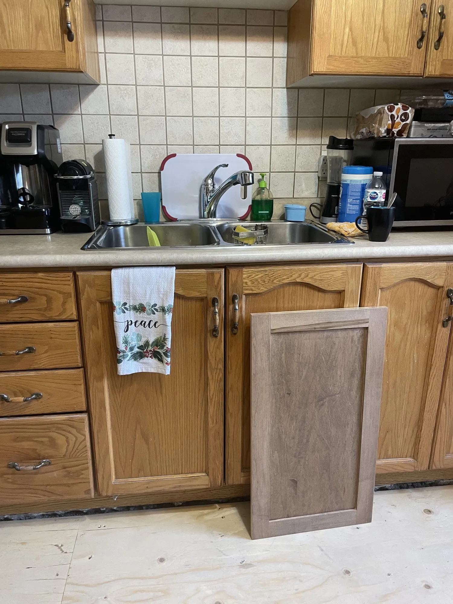 A grey-brown stained sample door standing against the original golden oak cabinets and tiled backsplash during the estimate visit