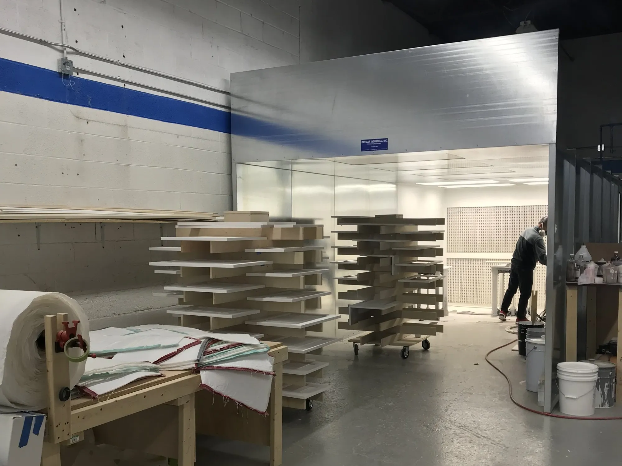 Doors and panels racked beside the spray booth, a sprayer working inside it