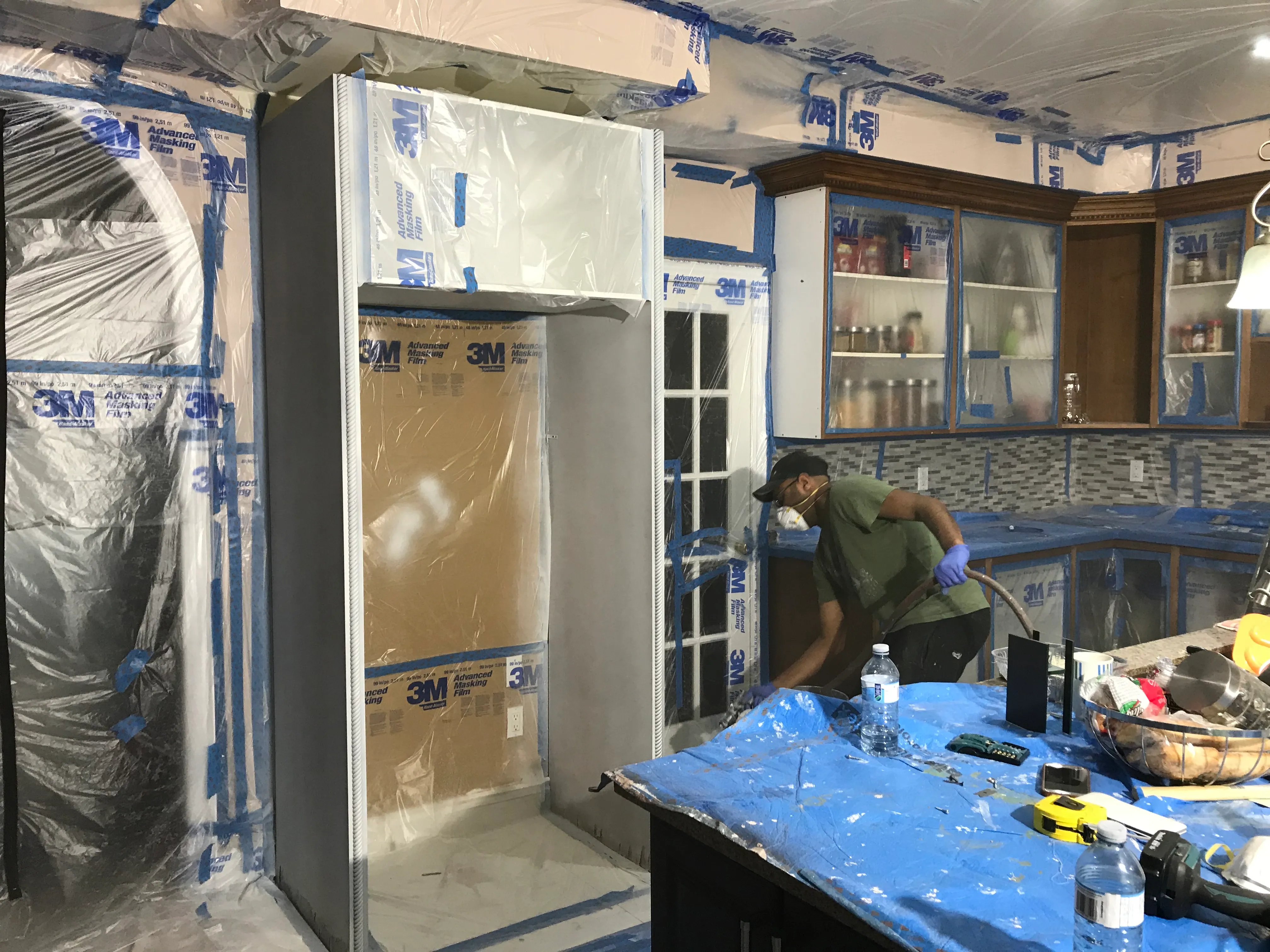 A technician in a respirator spraying inside a kitchen fully sealed in plastic sheeting and 3M tape