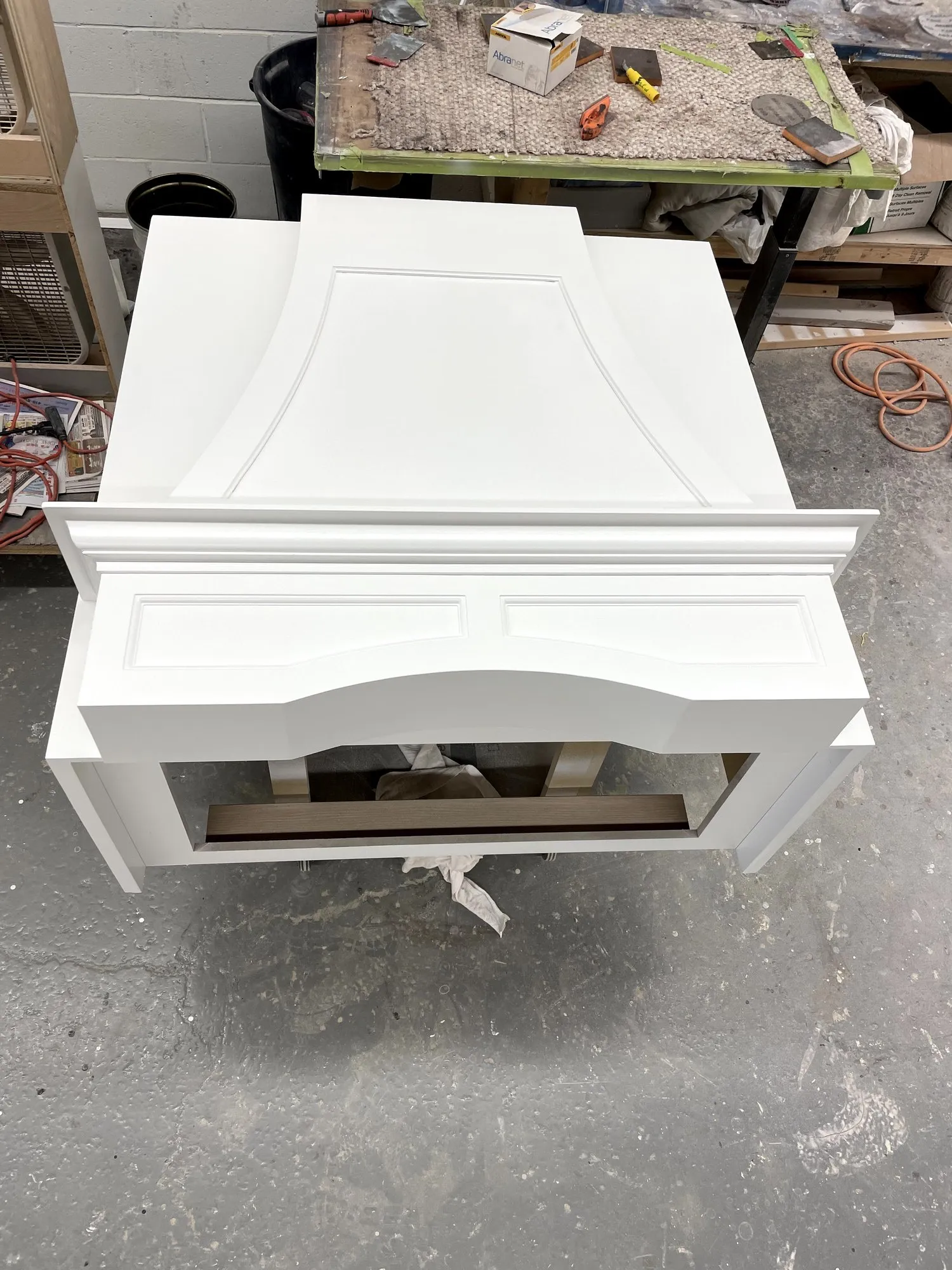 A range-hood surround on the shop floor, sprayed white, its curved apron and moulded panels finished before it goes back on the wall