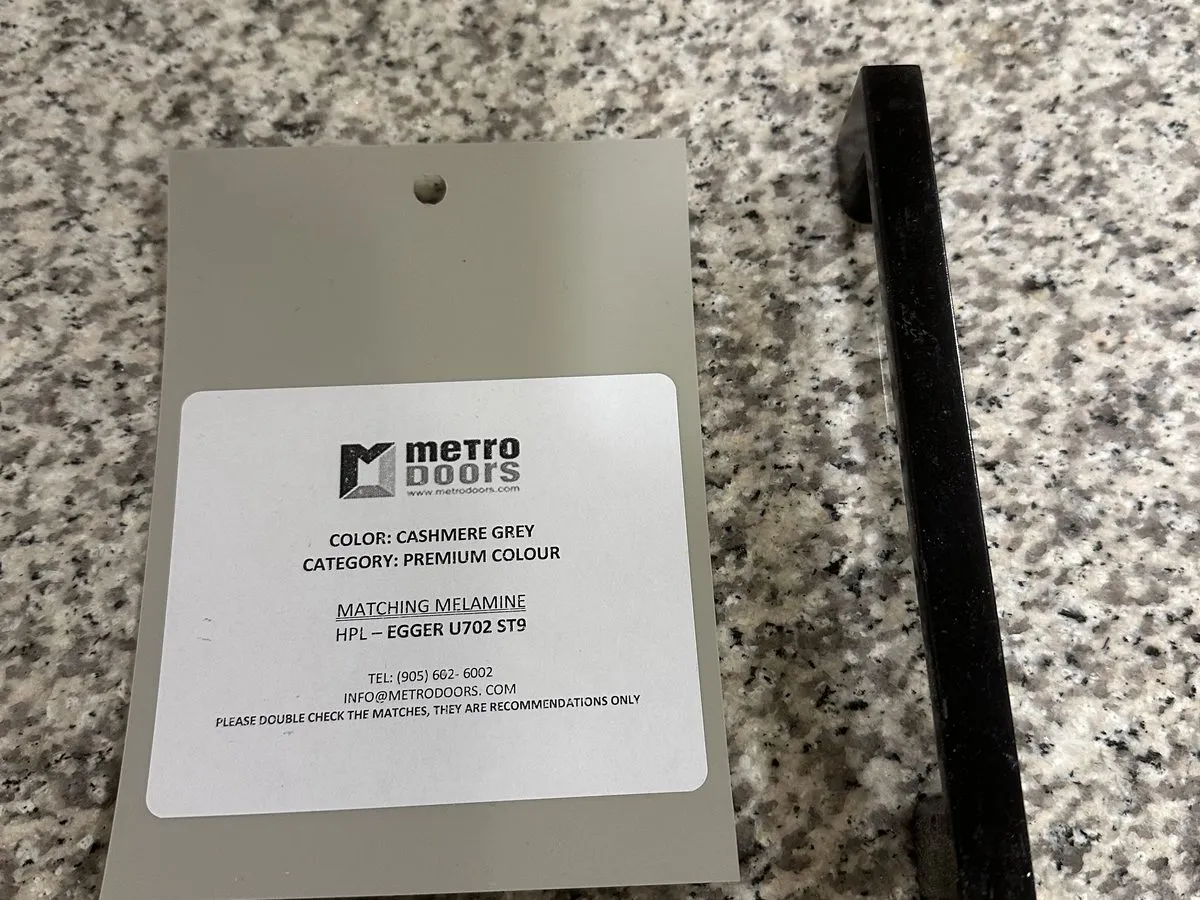 A door-material sample chip and a cabinet pull laid on a client's granite countertop, brought to their kitchen for a decision under their own light