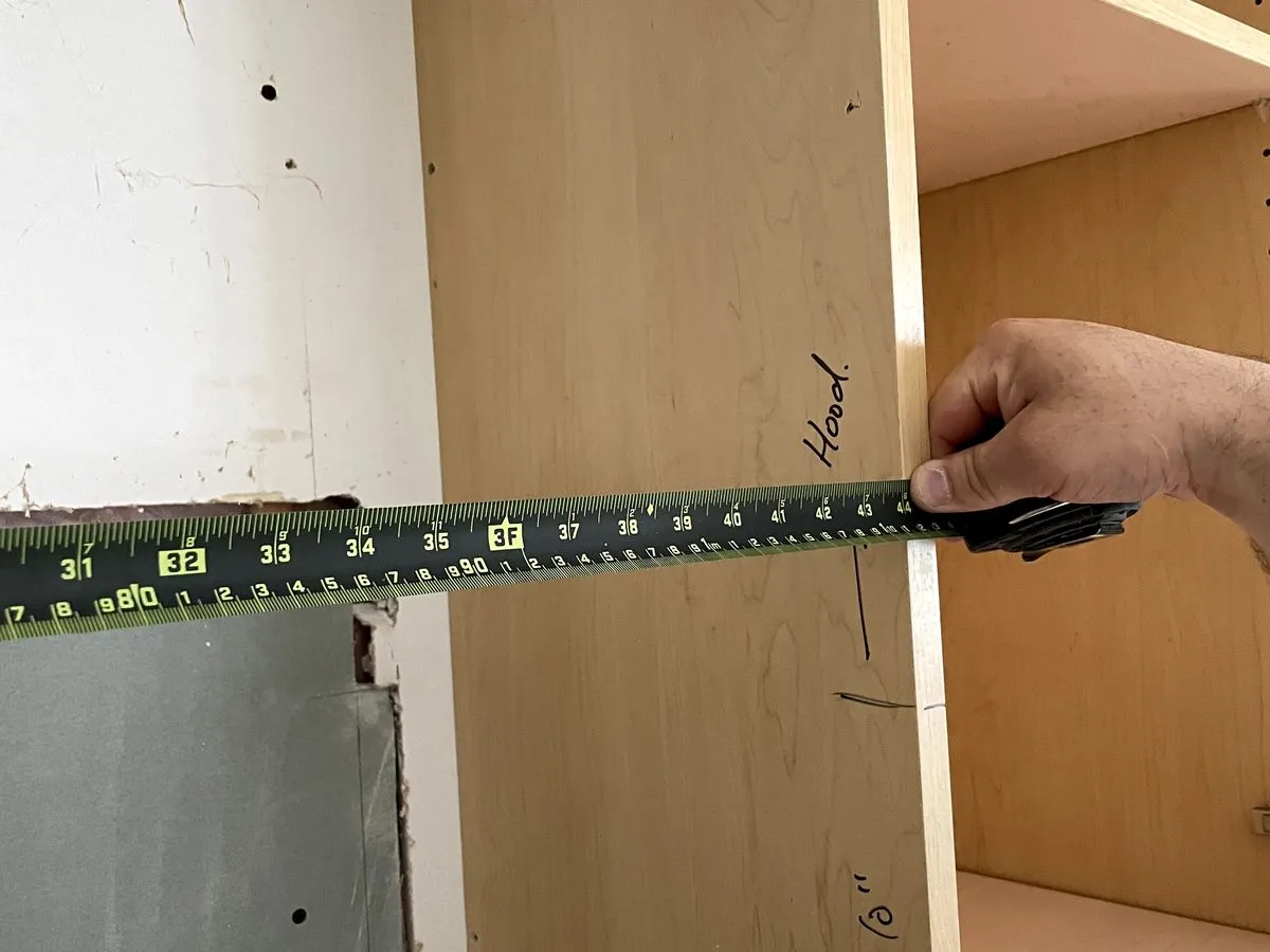 A hand measuring the inside of an open cabinet box with a tape measure, the opening marked in pencil