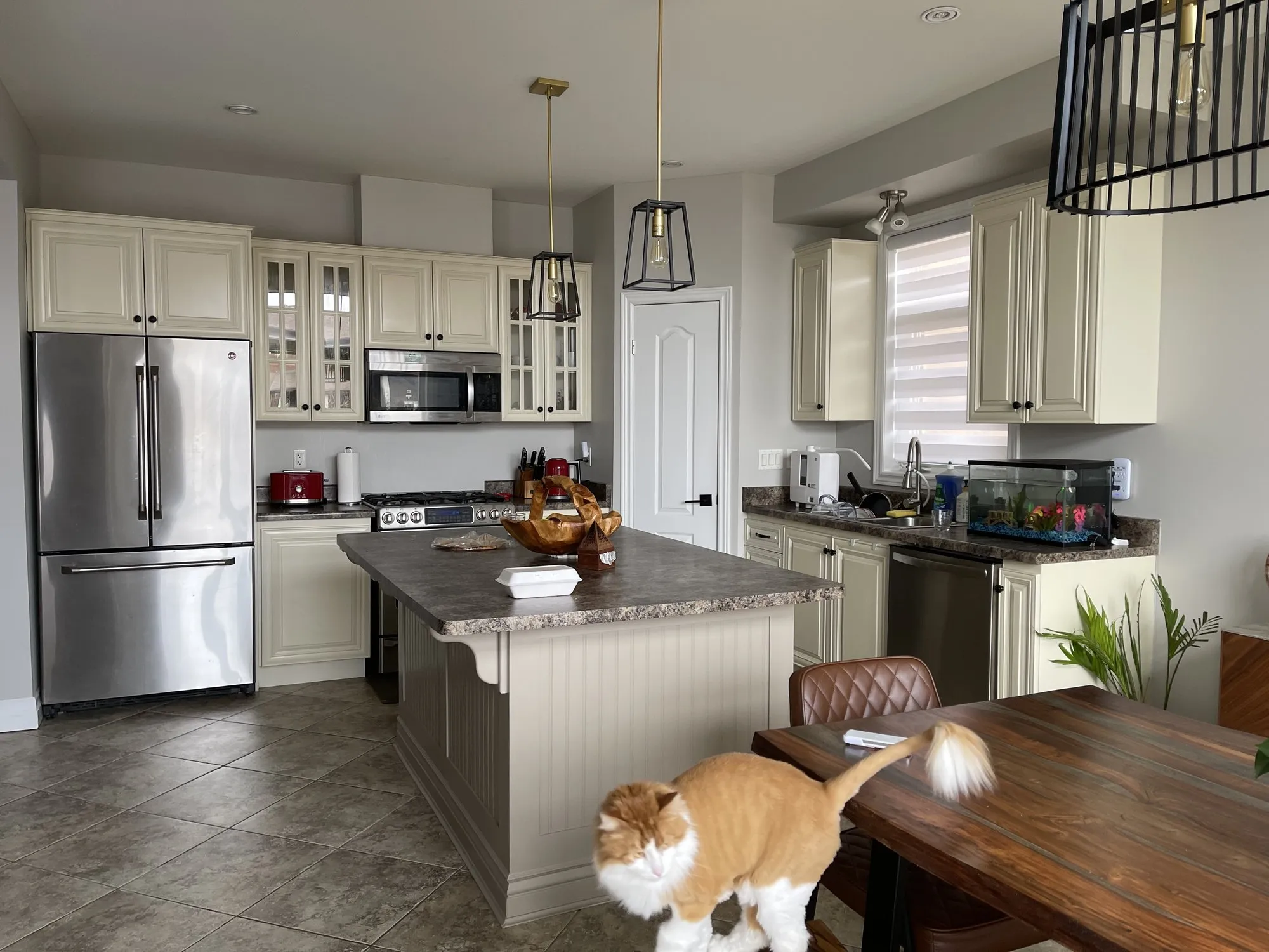 The original cream kitchen with glass-fronted upper cabinets, an arched panel door profile and a beadboard island, under speckled laminate counters