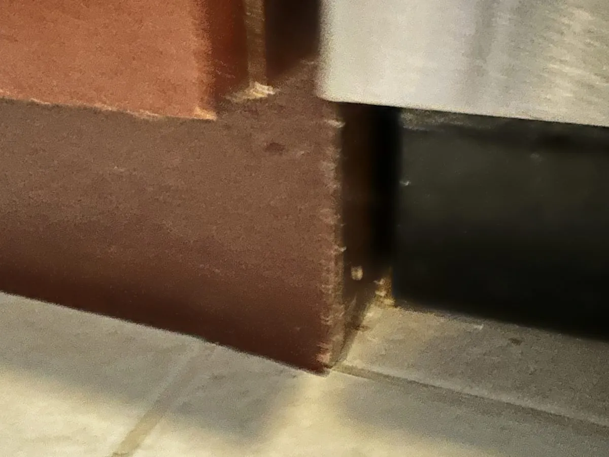 A close-up of a cabinet panel's finish separating from the substrate at a base corner, the failure visible edge-on