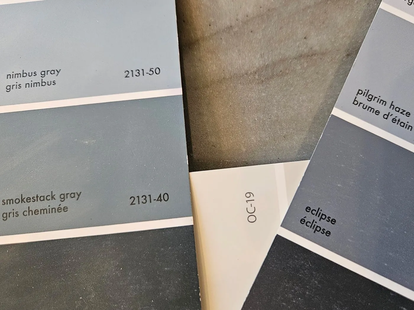 Benjamin Moore chips laid out on a customer's own countertop — nimbus gray, smokestack gray, pilgrim haze and eclipse, being compared against the stone they will sit beside