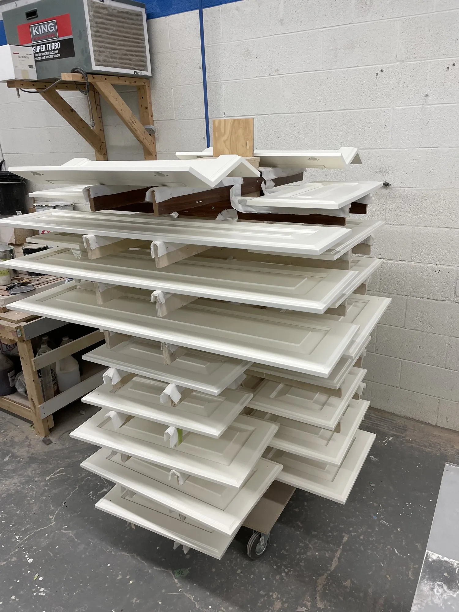 Freshly sprayed doors and raised panels stacked on a drying rack in the shop, each on its own tier so no two faces touch while the finish cures