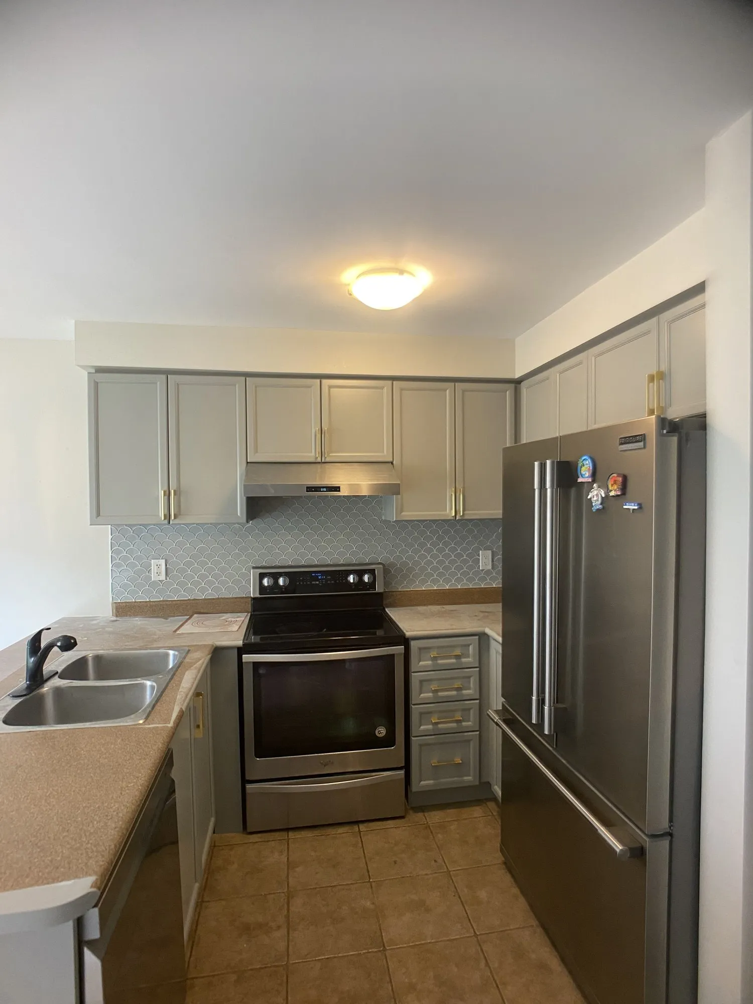 The same kitchen after refinishing, light grey with brass handles, the original beige counters and tile backsplash unchanged