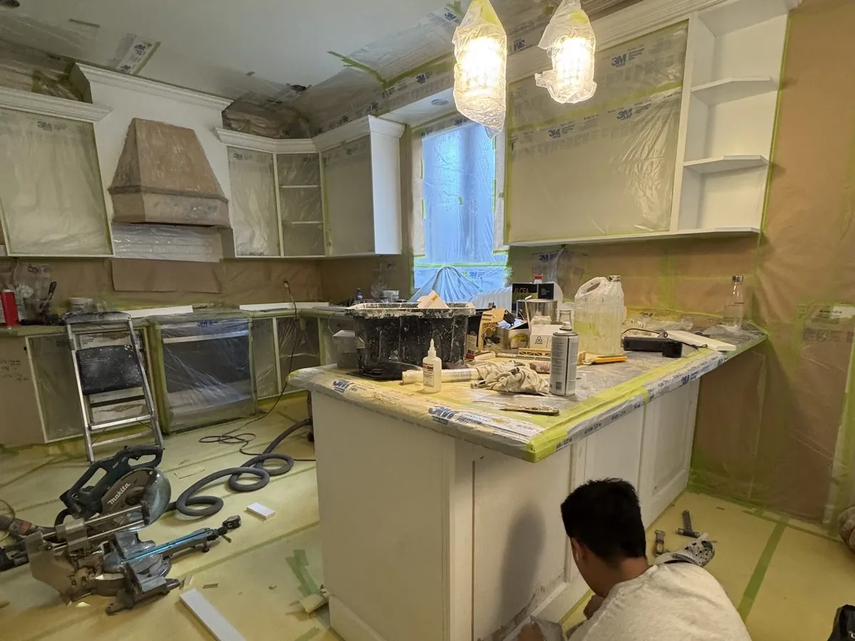 A kitchen fully sealed in plastic sheeting and tape, a crew member spraying cabinet frames behind the containment