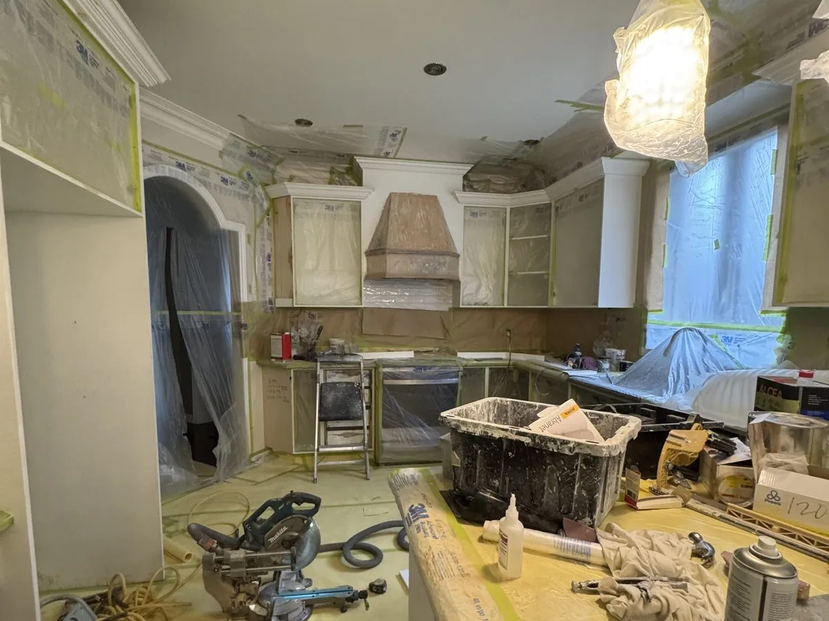 A kitchen sealed behind plastic sheeting and painter's tape mid-spray, crew visible at work inside the containment