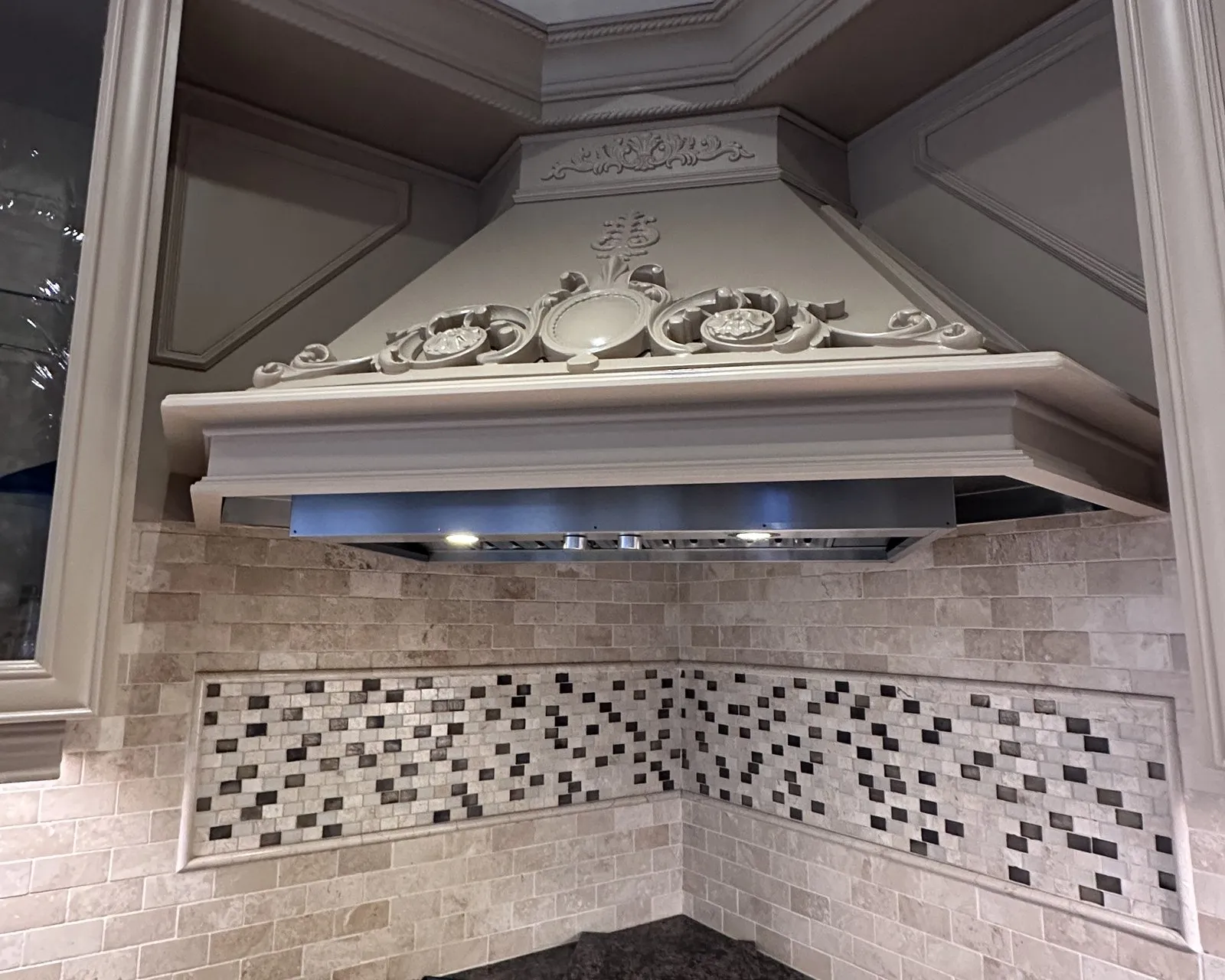 A carved wood range hood painted a soft warm grey, the moulded scrollwork and rosette detail picked out under the hood lighting